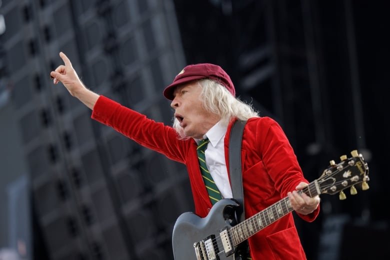 An older man with long, white hair wearing a red velvet jacket and red cap holds an electric guitar and raises one hand in the air as he shouts on a stage. 
