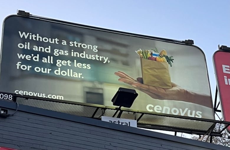 A billboard reads "Without a strong oil and gas industry we'd all get less for our dollar," and shows an outstretched hand holding a small paper bag of groceries.