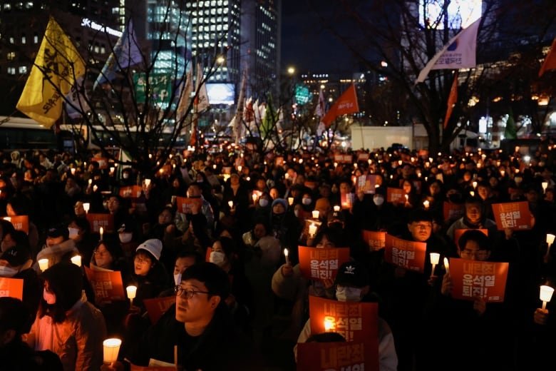 Hundreds of people are shown outdoors in a nighttime scene, holding small candles or signs.