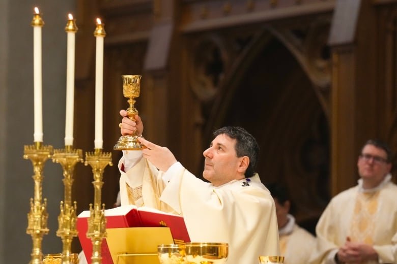 A bishop stands at a pulpit and holds aloft a golden cup. He is white, middle-aged and wearing white robes, looking up to the cup. There are 3 candles in the foreground, and a clergyman behind him.