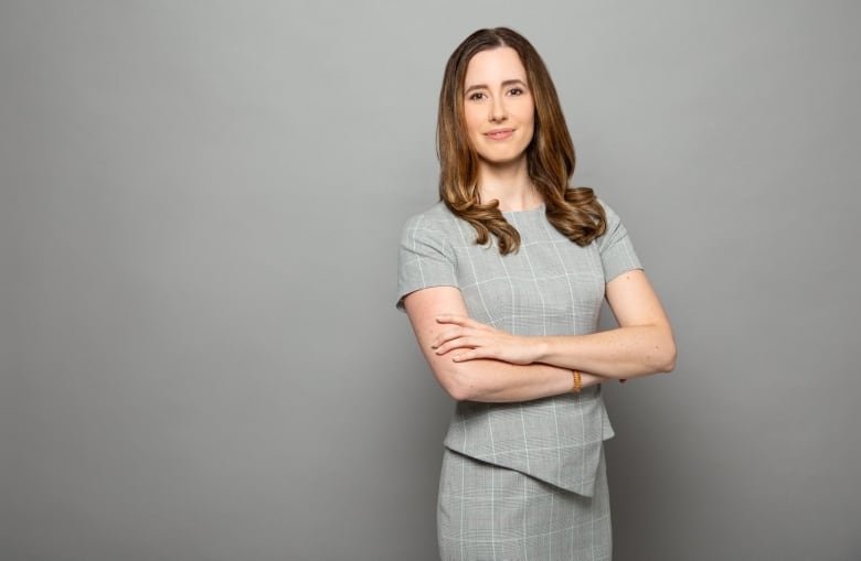 A slim white woman with long brownn hair n a grey dress with short sleeves stands with her arms folded looking at the camera.    
