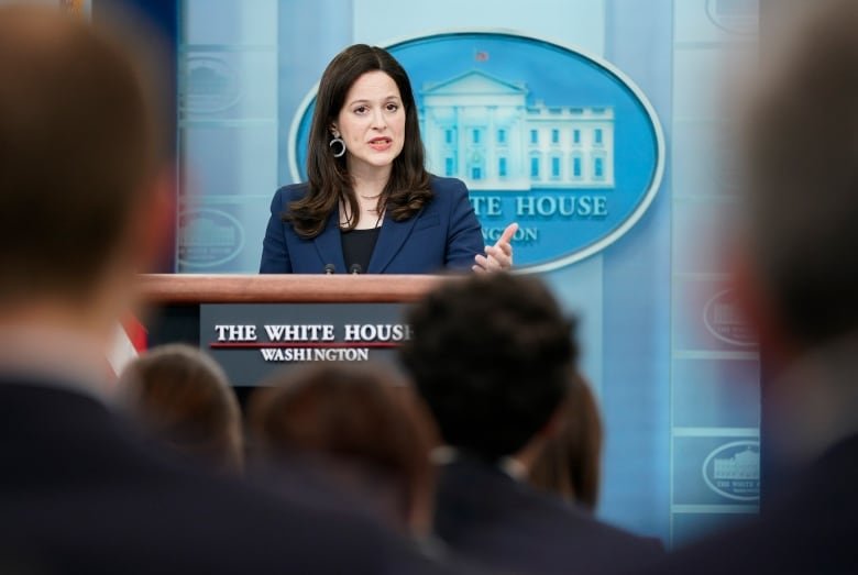 A woman with long, dark hair wearing a navy suit stands behind a podium. 