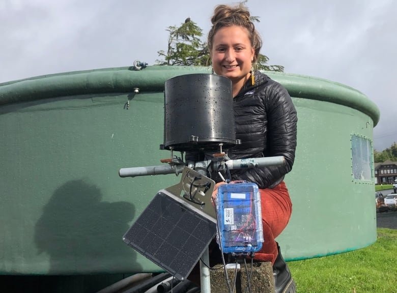 A woman with light brown hair, a black jacket and orange pants sits behind a black bucket to collect rainfall, with a circuit board attached.