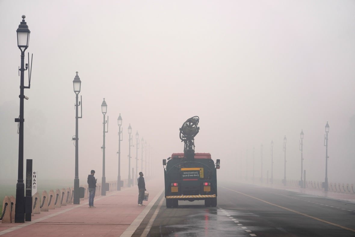 A truck drives through heavy smog along a nearly empty street. Two people stand near it on a sidewalk.
