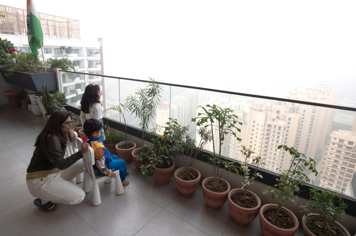 A woman crouches beside two children sitting on small chairs on a high-rise balcony. Beyond the balcony’s clear railing, heavy smog almost obliterates the city skyline.