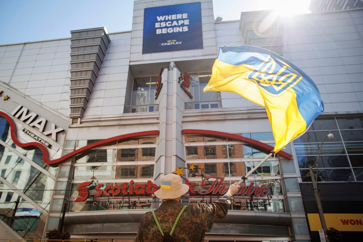 A man wearing military fatigues waves a red and blue flag in front of a movie theatre.