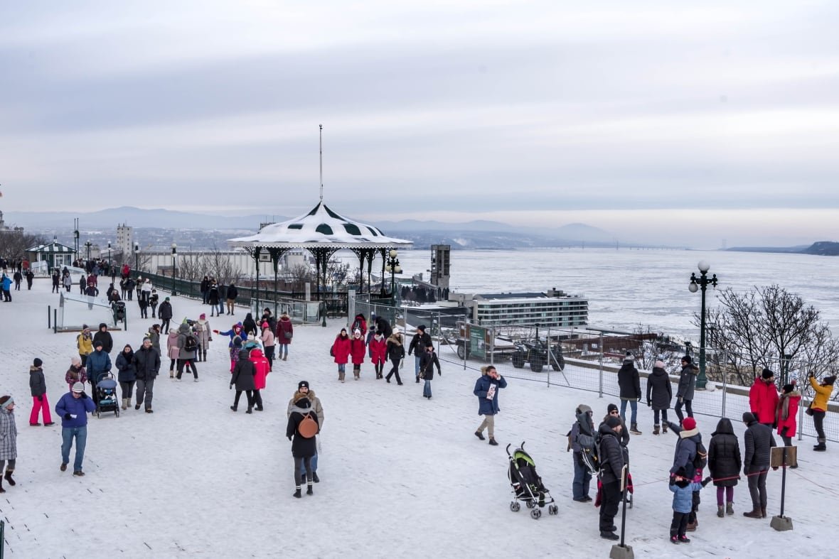 People walk on a snowy terrace with a river and mountains in the background.