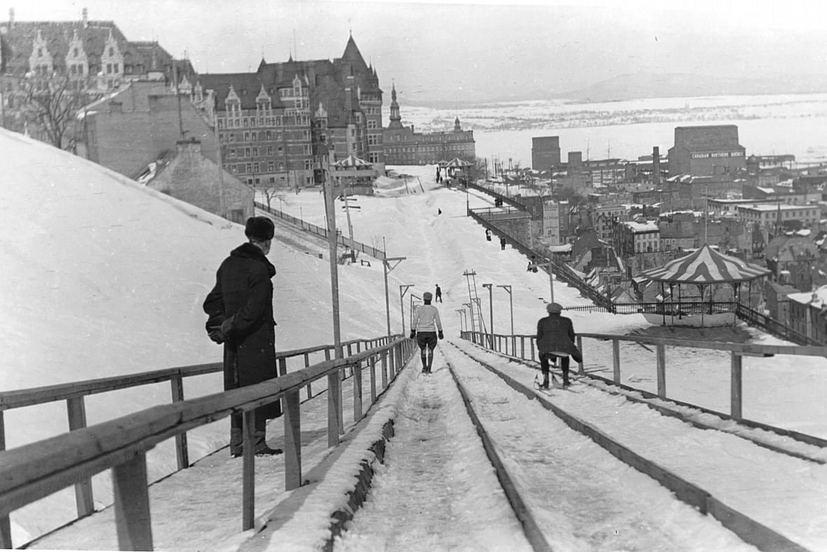 black and white photograph of a man watching two people slide down an ice slide. 