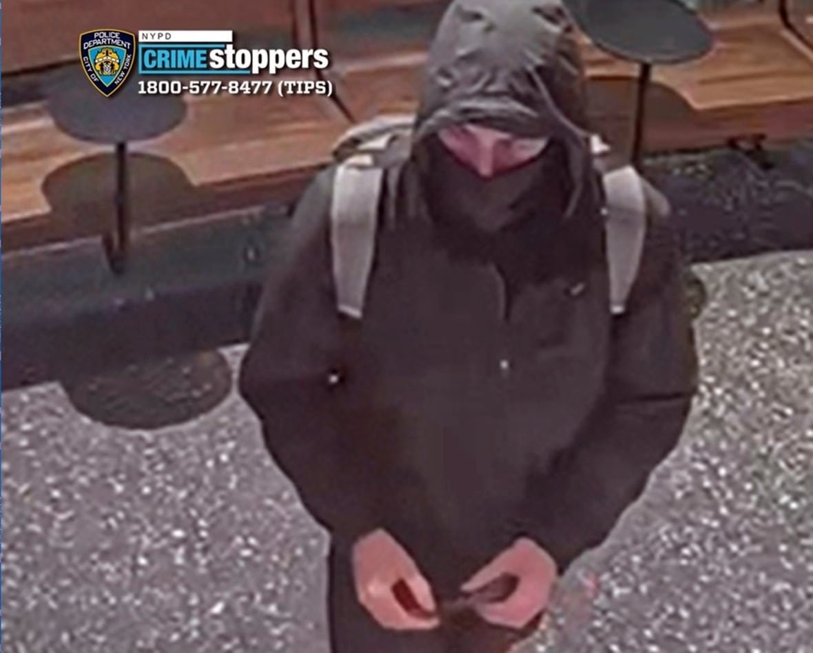 A man in a black hoodie with a black mask covering the lower half of his face is seen in a photo. He is wearing a light grey backpack.