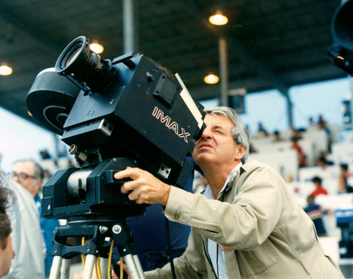 A man with grey hair looks through the viewfinder of an Imax camera pointed up at the sky. 