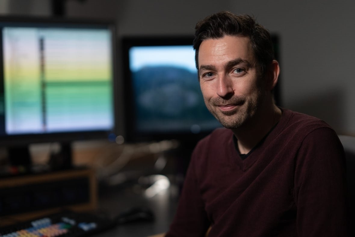 A smiling man is seated in front of two computer screens. 