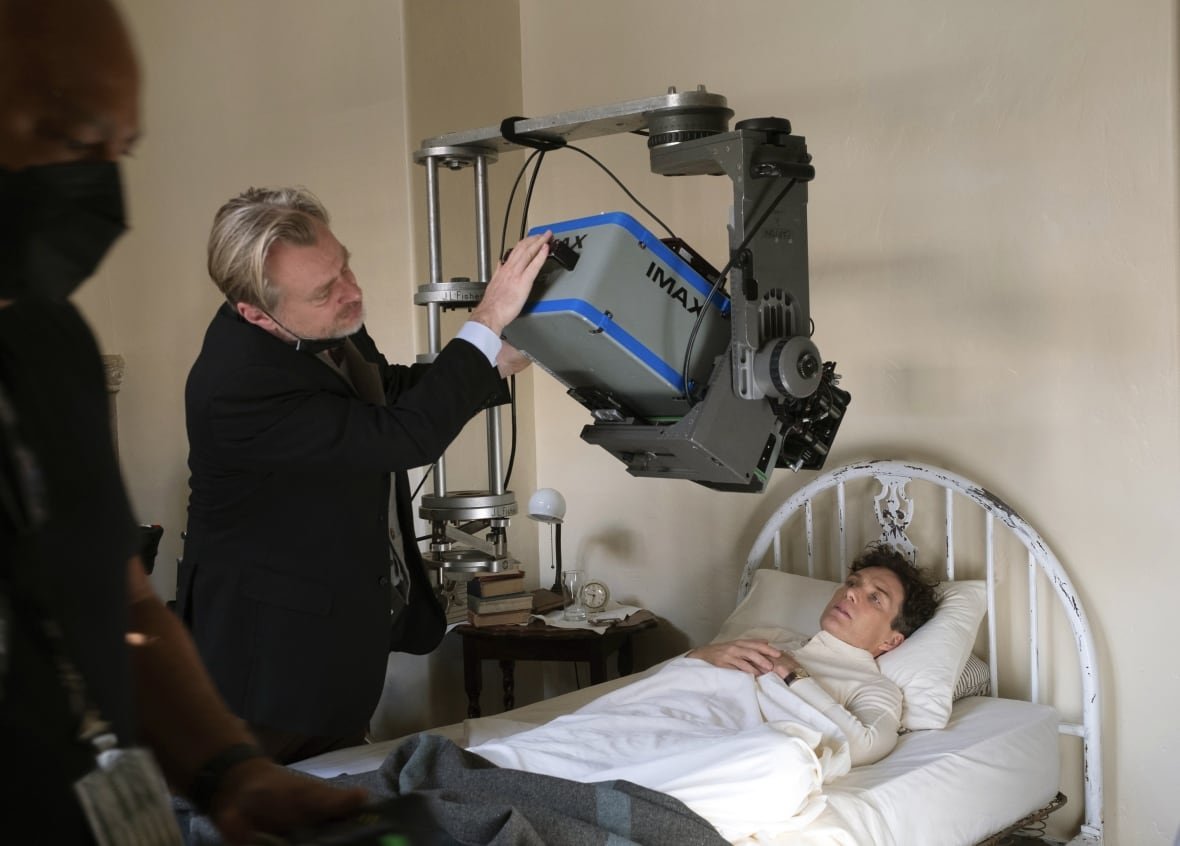 A blond man in a dark suit jacket adjusts an Imax camera hanging over a thin man in a bed. 