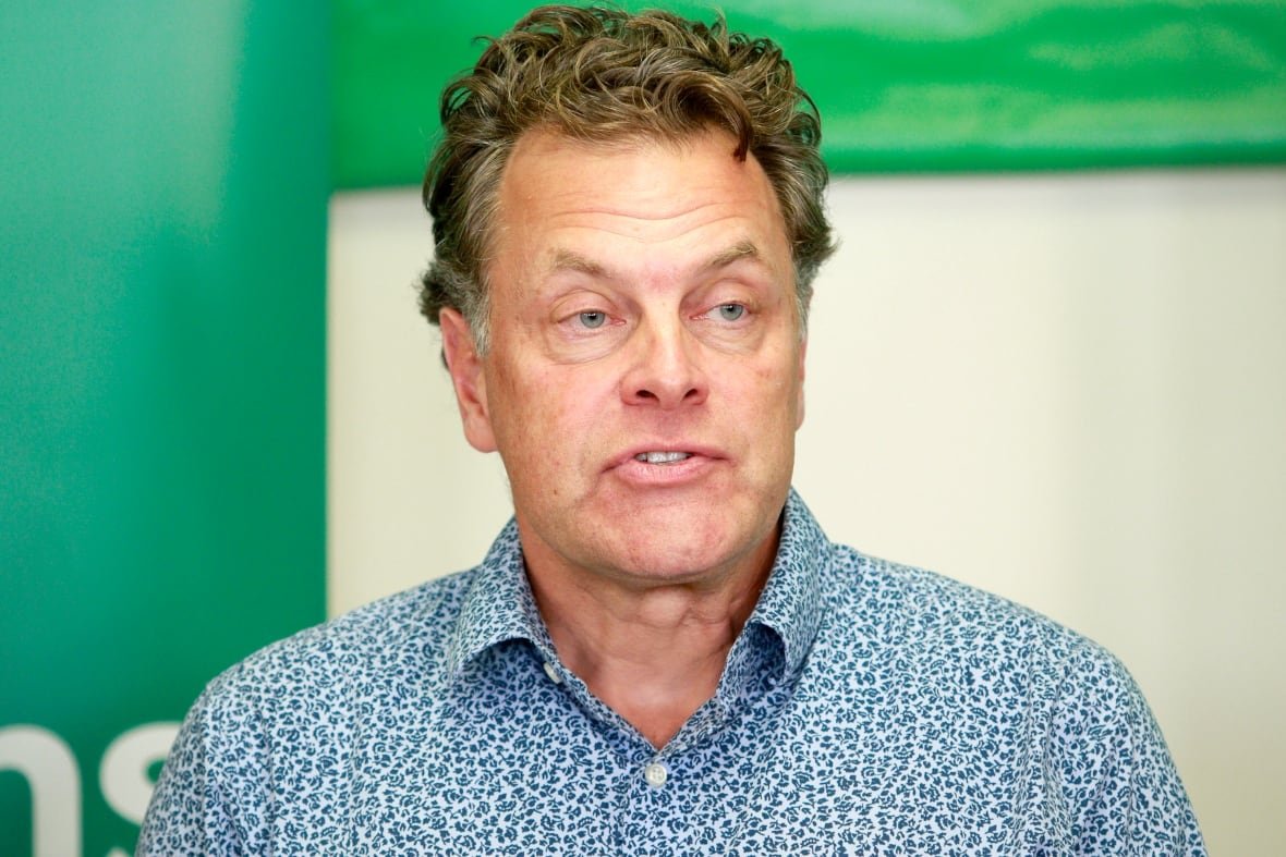 A man wearing a blue patterned shirt speaks in front of a green background.