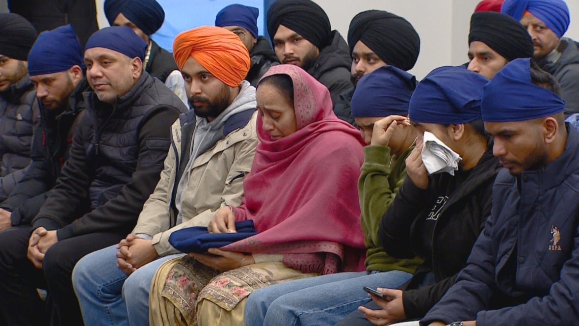 People attend the funeral of Harshandeep Singh. They are seated in a row, some are crying. The woman in the middle of the photo is wearing a pink head scarf and others in the row are wearing turbans and other head coverings. 