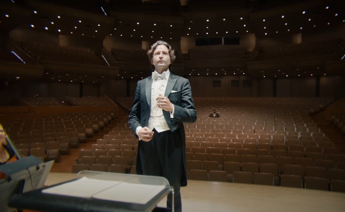 A man wearing a tuxedo stands on a stage facing away from an auditorium. In the auditorium one man can be seen seated.