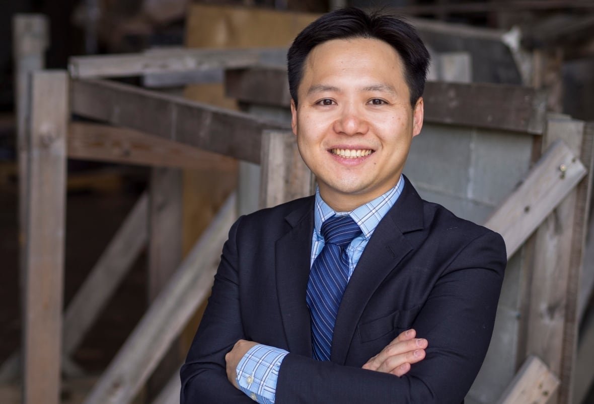 An Asian man in a blue suit jacket, blue shirt and blue tie stands and smiles in front of a stack of wood and stone materials.    