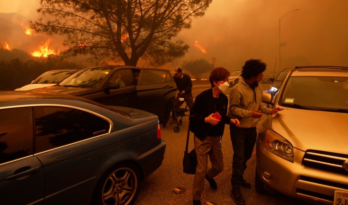Two people walk between cars, one wearing a mask. Behind them, a man is in the middle of pulling something out of the backseat of his car, reaching into the open car door. The road appears filled with cars, and the entire image is cast over with a hazy orange. In the distance, off the road, flames are visible. 