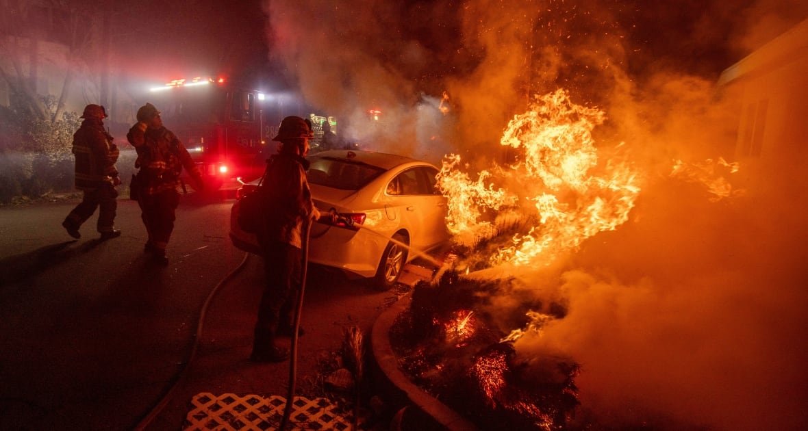 Firefights spray water on a fire. A car is parked near the firefighter with the hose, and nearby there are more firefighters. 