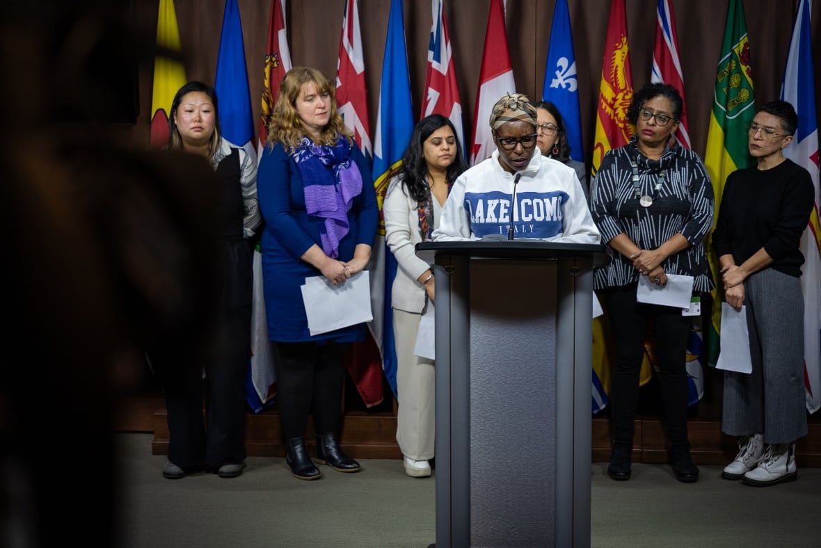 A woman wearing a hoodie and a head scarf speaks at a podium flanked by six other women.
