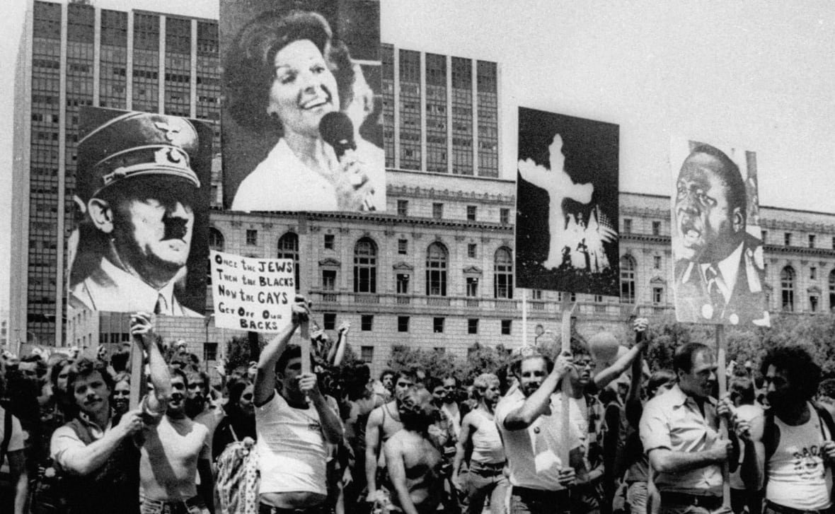 A crowd of people protest in a black and white photo. They hold signs of four faces up above them. Another sign says "Once the Jews. Then the Blacks. Now the Gays. Get Off Our Backs". 