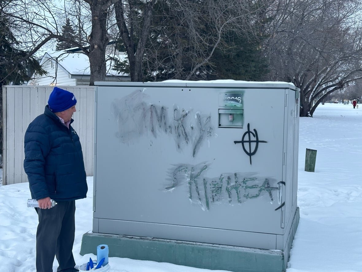 A man stands in front of some graffiti outdoors that's been partially deleted, with a swastika, the words "M.K.Y." and "Skinhead" still legible.
