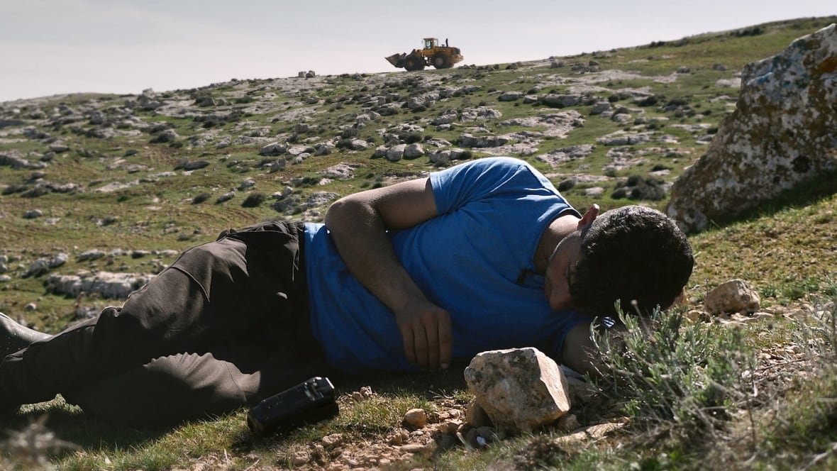 A man lies on the ground on a grassy hill studded with rocks. A bulldozer is visible at the top of the hill beyond him. 