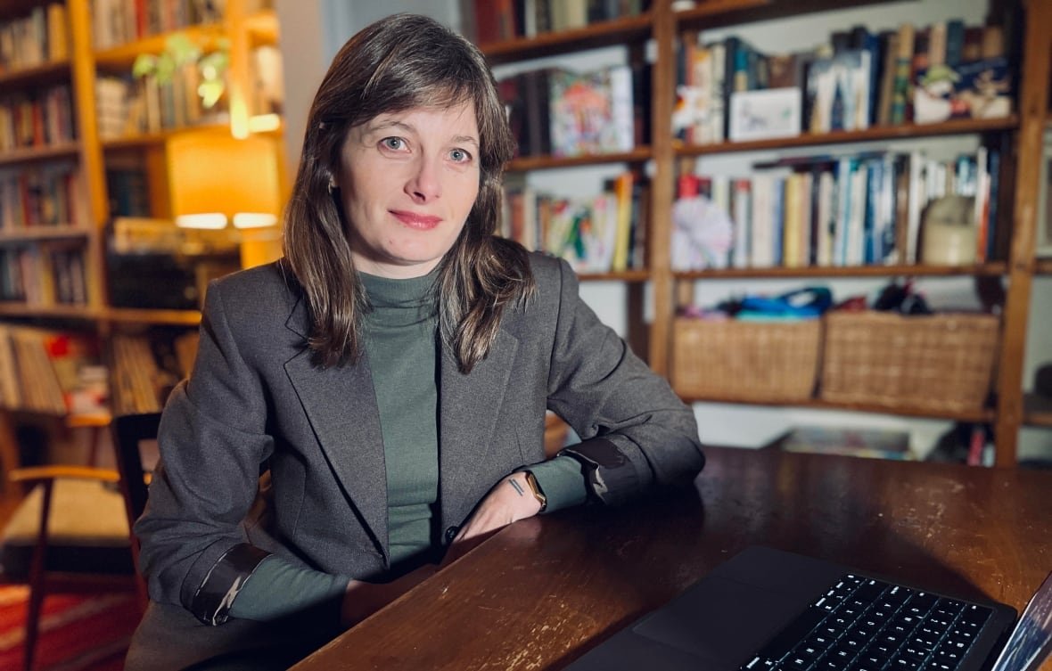 A woman sits at a desk with a laptop in front of her, smiling at the camera. She is wearing a blazer and an office with bookshelves is visible behind her. 