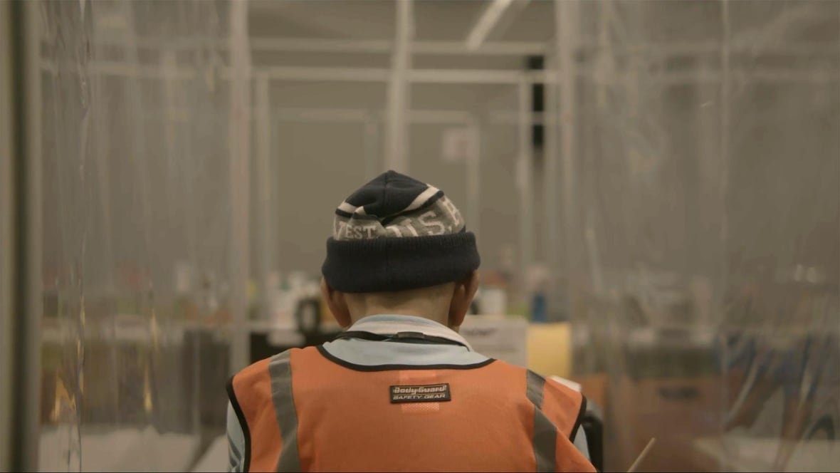 A person is seen from behind from the shoulders up. They are wearing an orange vest and a black and grey hat. All around them there are what appear to be plastic walls or dividers. 
