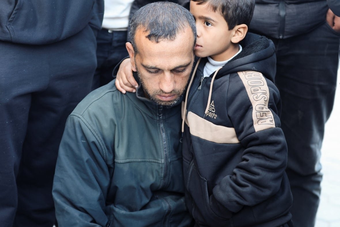 A boy embraces an older man, kissing the side of his head. Both look sad, and are in dark clothing. 