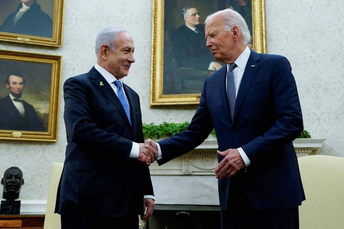 Two men in dark suits shake hands, smiling. 