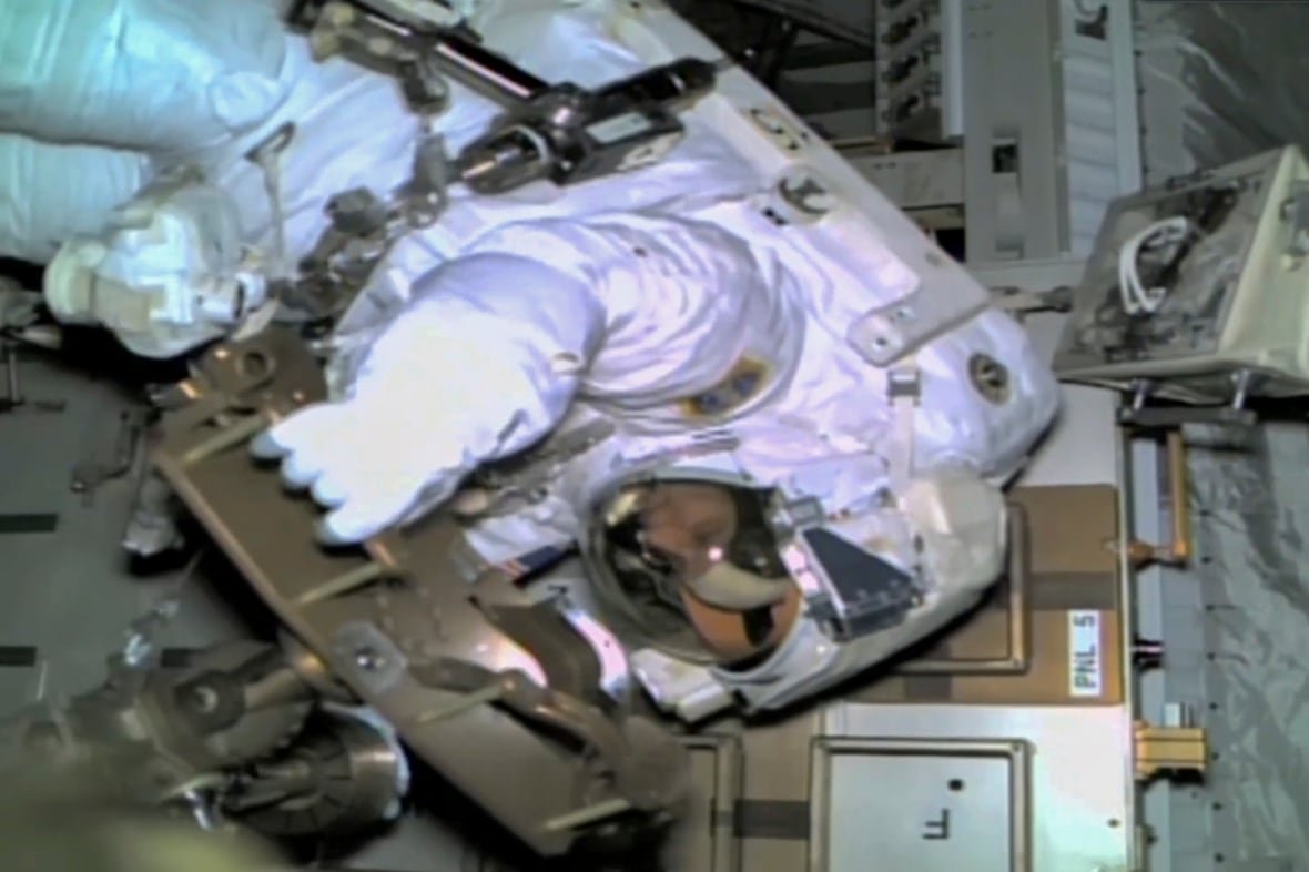 An astronaut floats seemingly upside down, back at the top of the image, facing to the left. A bunch of equipment is behind them and the edges of the photo darken beyond the flashlight aimed at the astronaut.