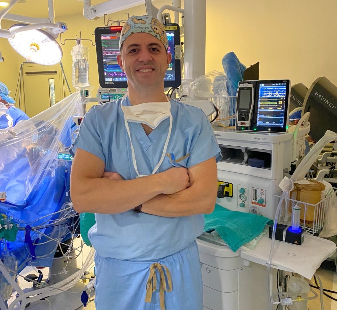A man in medical scrubs stands in front of anesthesia equipment in a hospital procedure room.