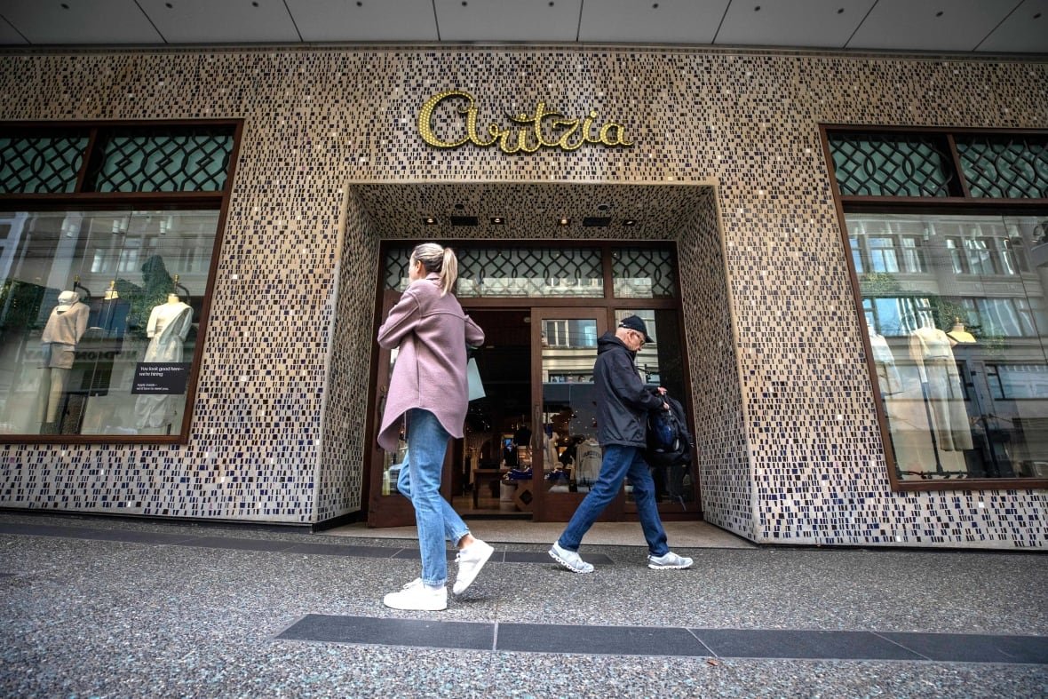 Two people walk in opposite directions in front of a store. The word "Aritzia" appears over its wooden doors in gold metal script.