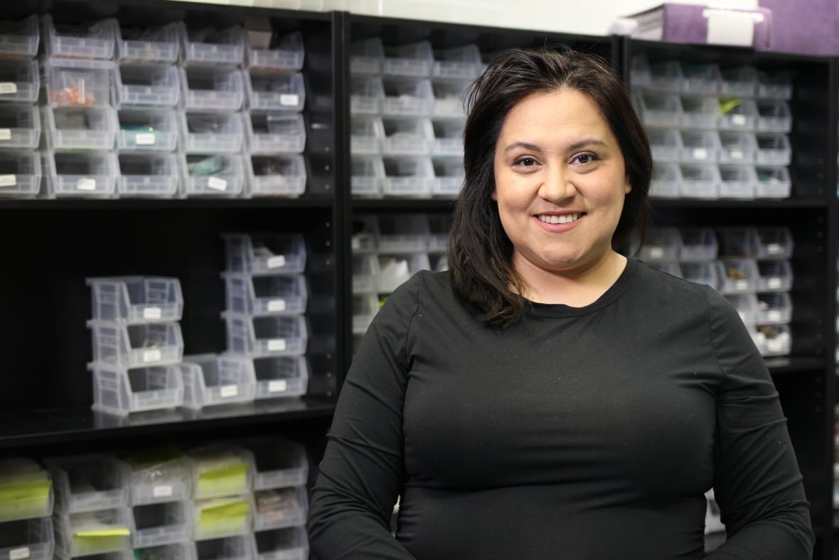 A woman in a black, long-sleeved t-shirt poses in front of a wall full of jewelry supplies.