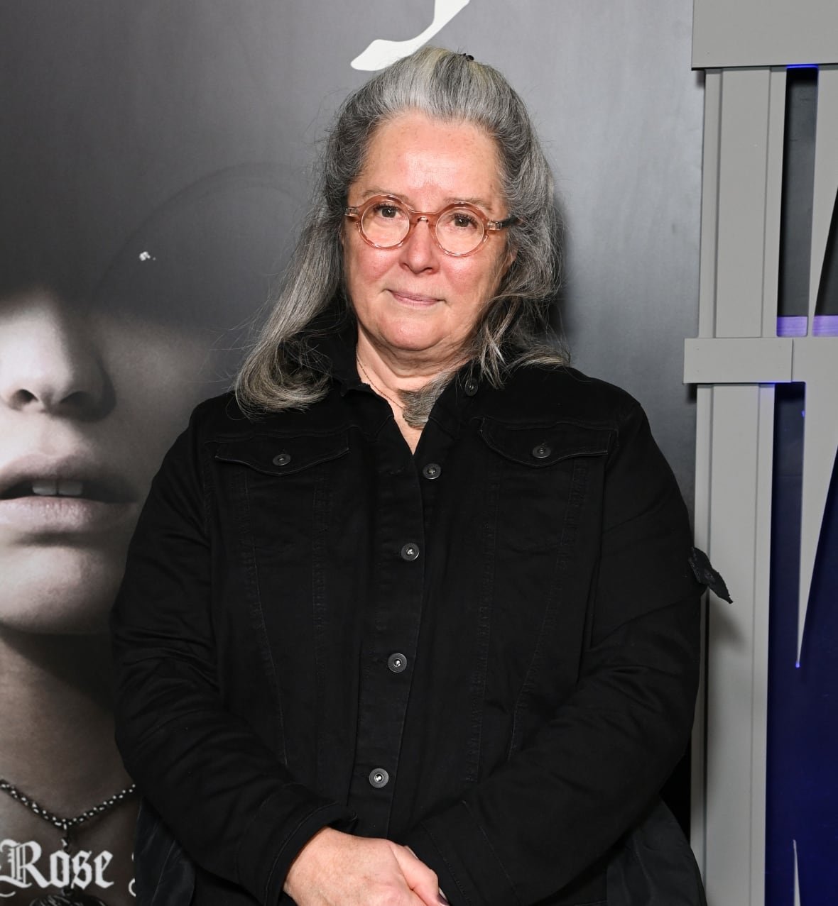 A woman stands with her hands folded in front of her, her head slightly tilted. She has grey hair and is wearing black and is standing in front of a grey backdrop. 