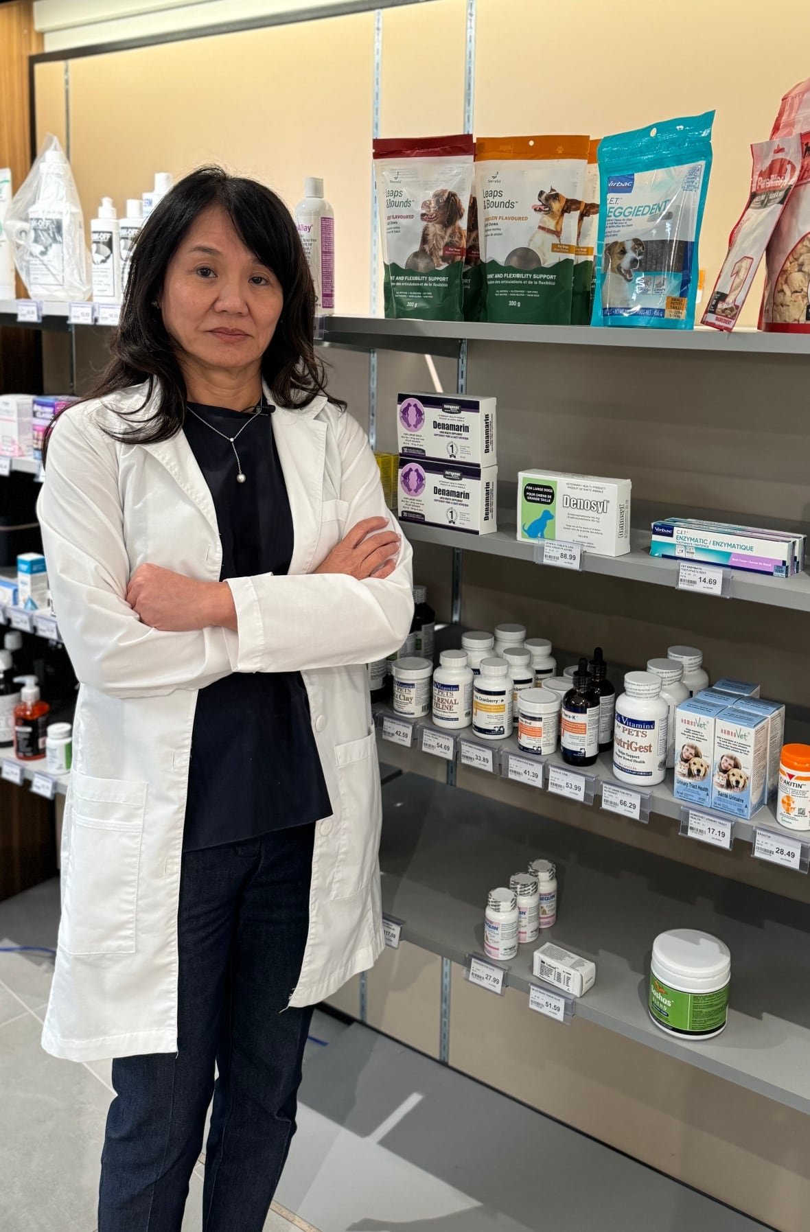 A female pharmacist stands in a white overcoat in front of a shelves filled with animal medication and pet food. 
