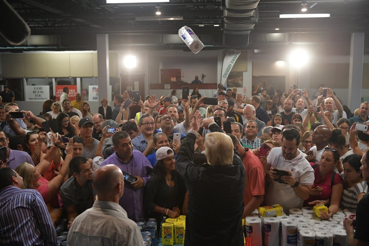 A paper towel package is shown high in the air above a crowd of dozens of people indoors. It appears to have been tossed by a man with his back to the camera.