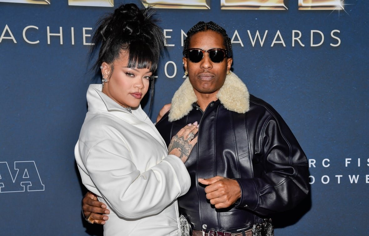 A couple pose in this photo. The woman, whose hair is done in a spiky up-do, is turned to the side and has her hand on the man's chest. The man, wearing a black leather jacket with a fuzzy collar, has his hand on the woman's waist. 