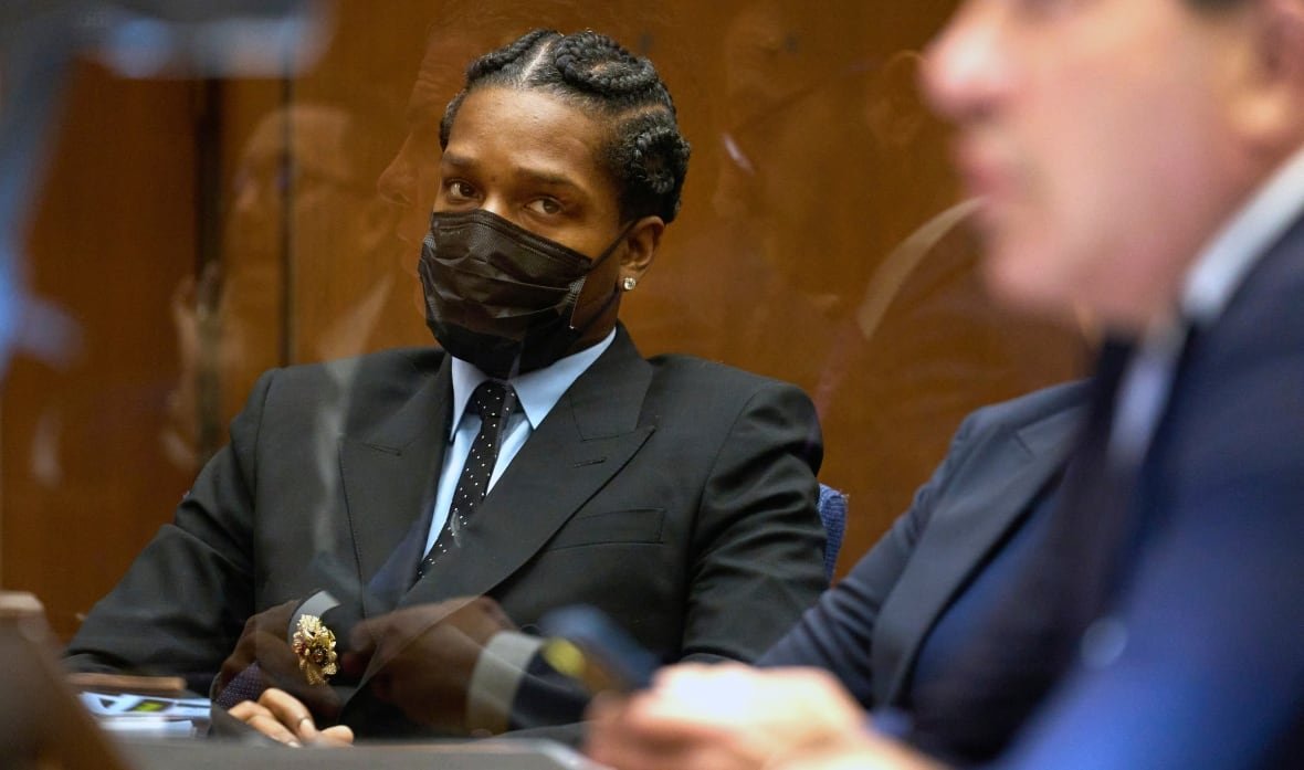 A man in a suit and wearing a black face mask is seen sitting in court. He is looking at a man in the foreground, who is in the middle of speaking, and is out of focus. 