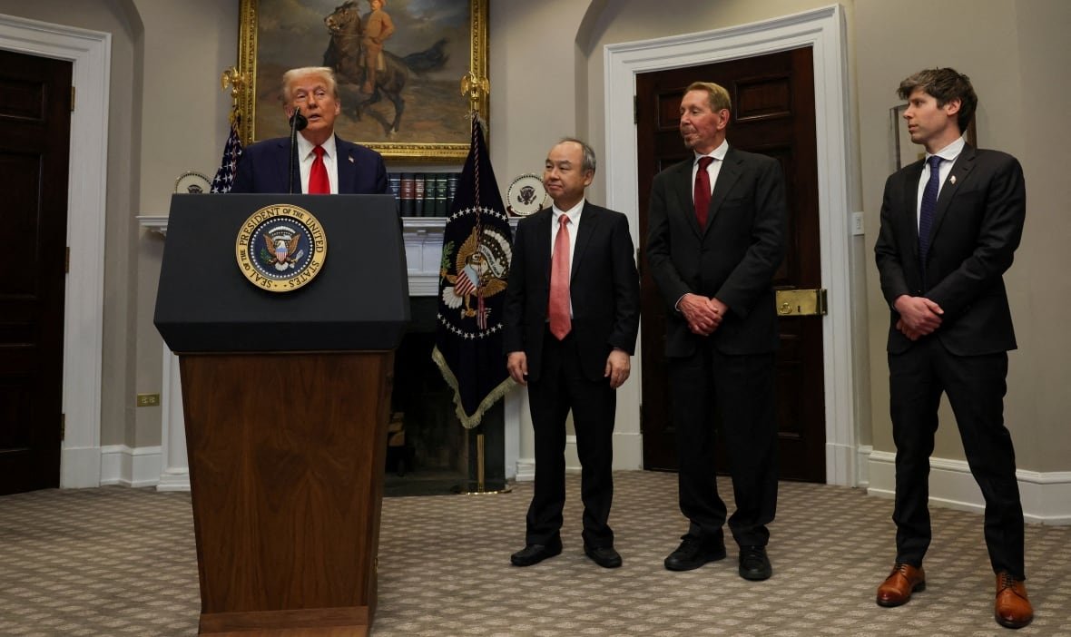 A man stands at a podium in the middle of a speech in a room. Three other men stand off to the right side of the image, looking at him. All are wearing suits. 