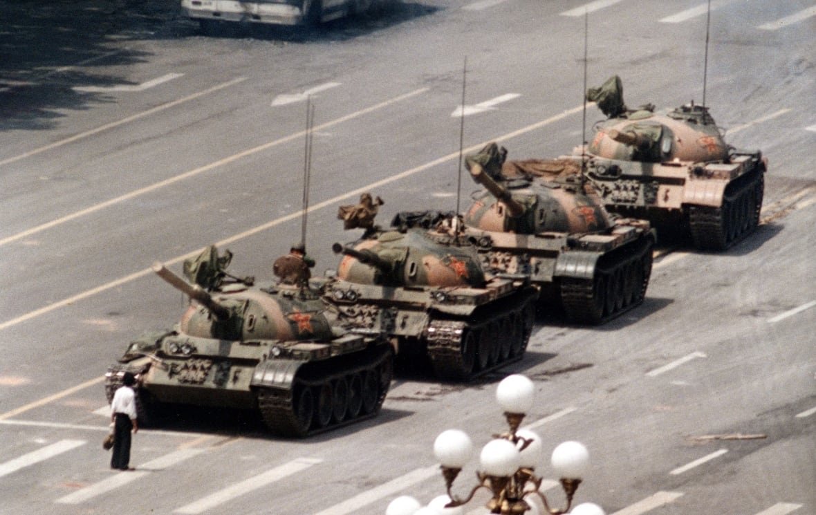 A lone man stands in the way of a column of tanks on a wide city road. 