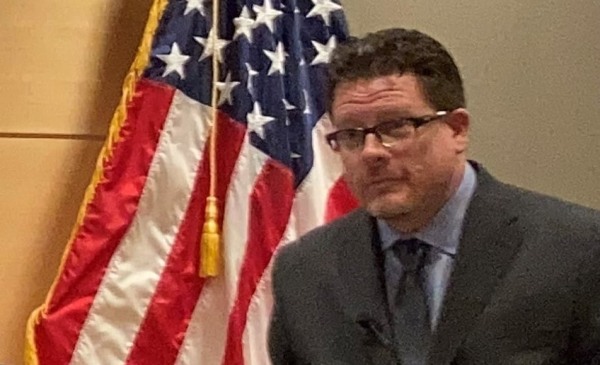 A man sits in front of a U.S. flag.