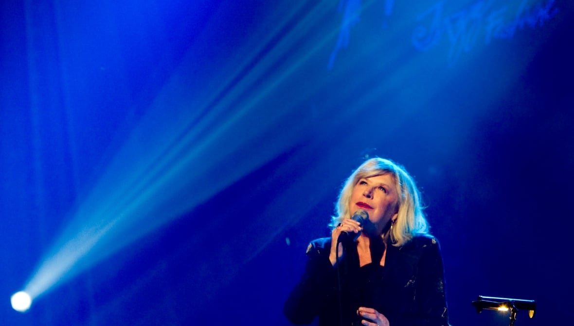 A woman sings into a microphone in front of a blue backdrop. A spray of stage lights shines from the bottom left corner of the image up diagonally over her head. 