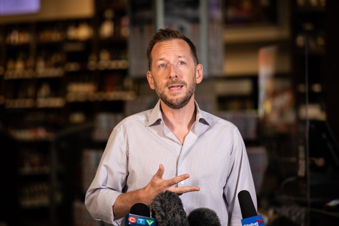 A man speaks in front of a cluster of microphones. The interior of a store is visible in the background. 