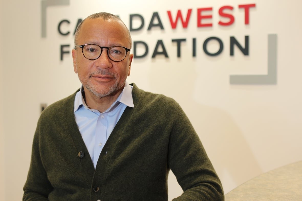 A man wearing glasses, a blue button-down shirt and a green cardigan poses in front of a wall bearing the logo of the Canada West Foundation.