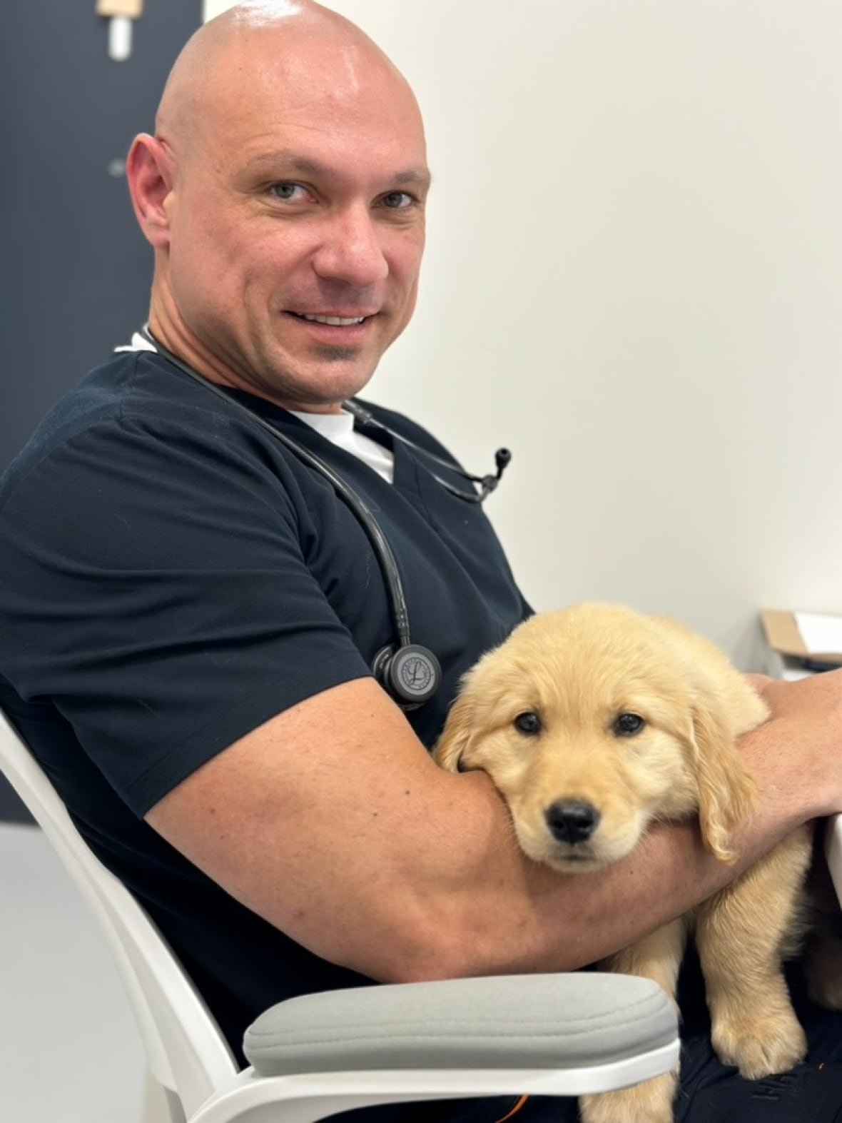 A man in medical attire smiles at the camera, holding a golden retriever puppy.