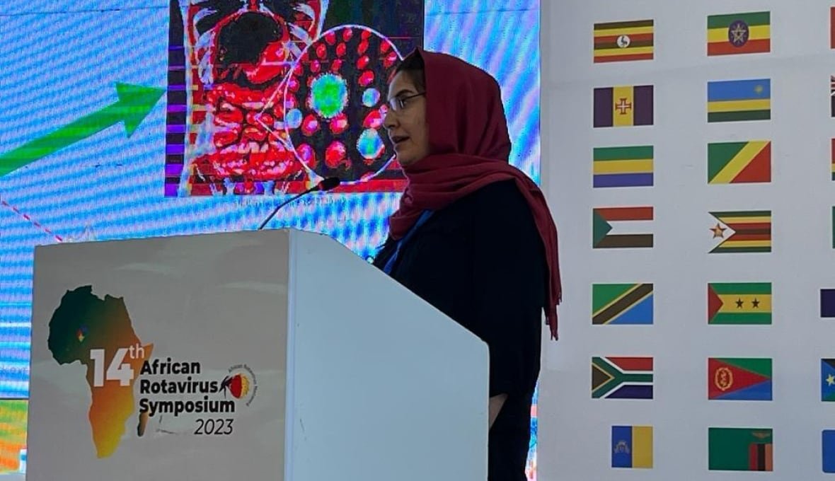 A woman in a red headscarf stands at a lecturn speaking in front of a projected slide presentation. 