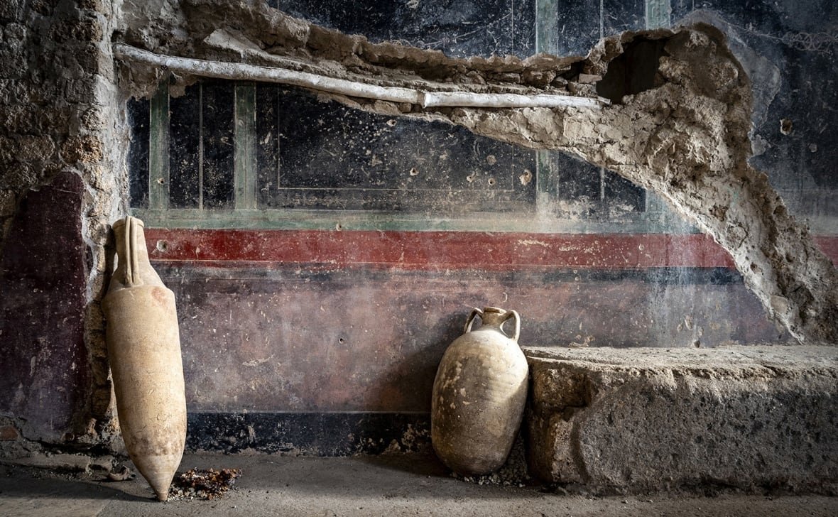 Two jars lean against a dusty wall covered in smudged red and black stripes for decoration. There is a large chunk of the wall that has been damaged and an oblong shape, perhaps a pipe, runs along the line of the damaged, exposed section. 