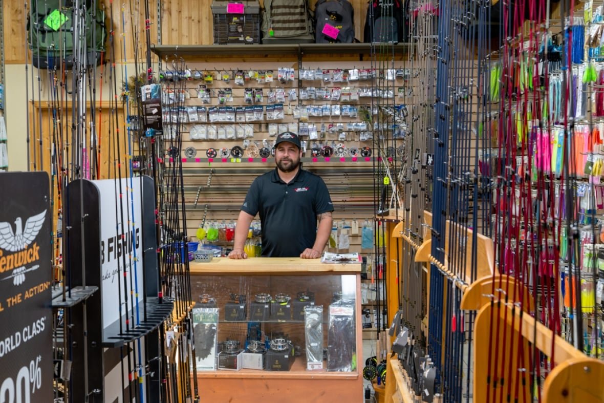 A man stands behind the counter of a store lined with fishing supplies. 