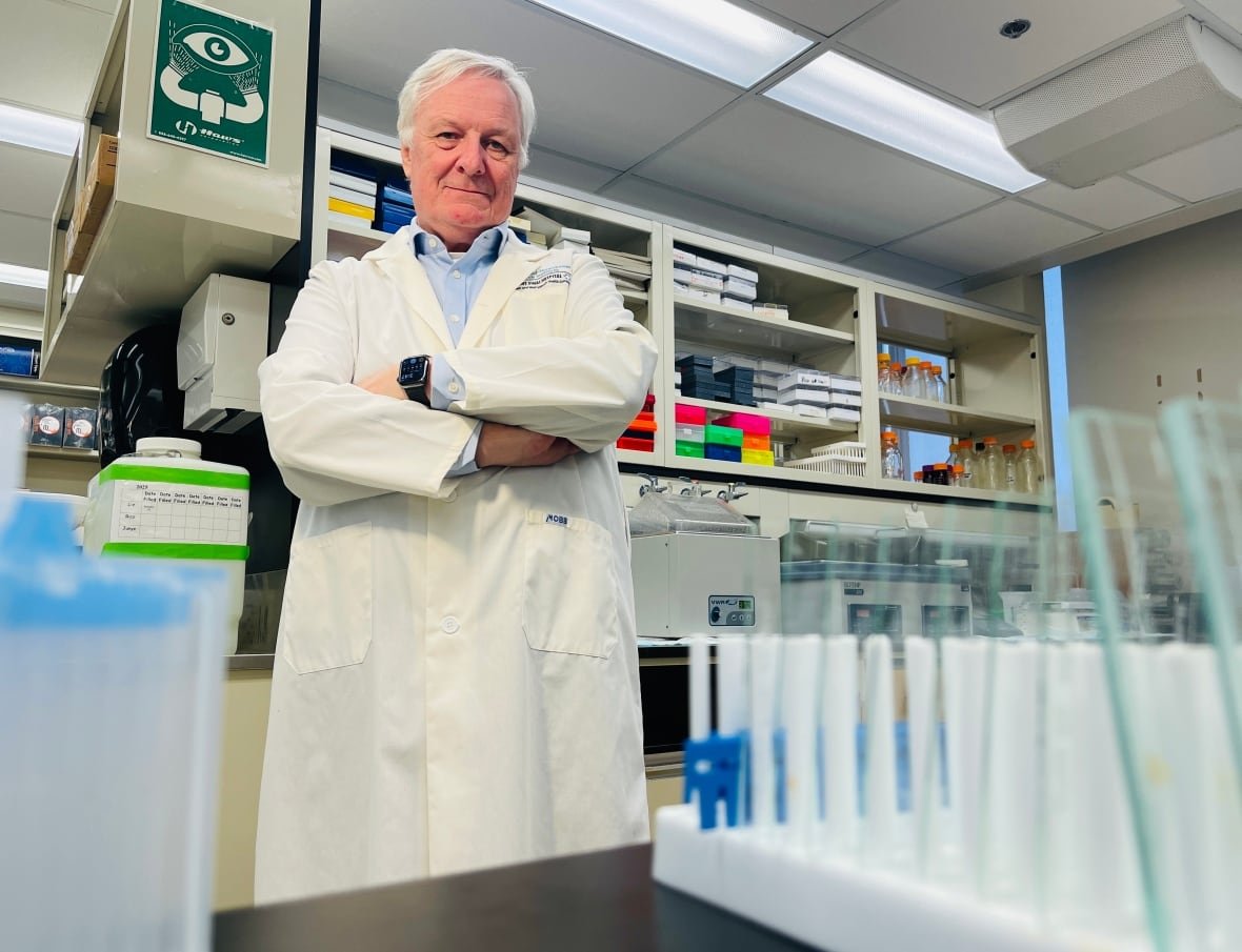 Man with white hair wearing a lab coat standing in a lab behind test tubes. 